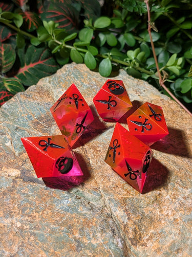 Red and pink polyhedral dice with black symbols on a stone surface with green foliage in the background